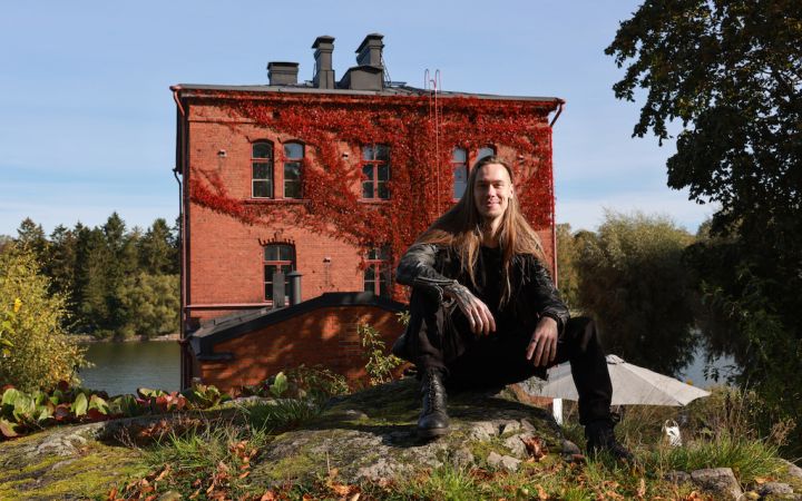 Tiilinen Venetsia-talo ja edustalla pitkähiuksinen istuva, hymyilevä mies.