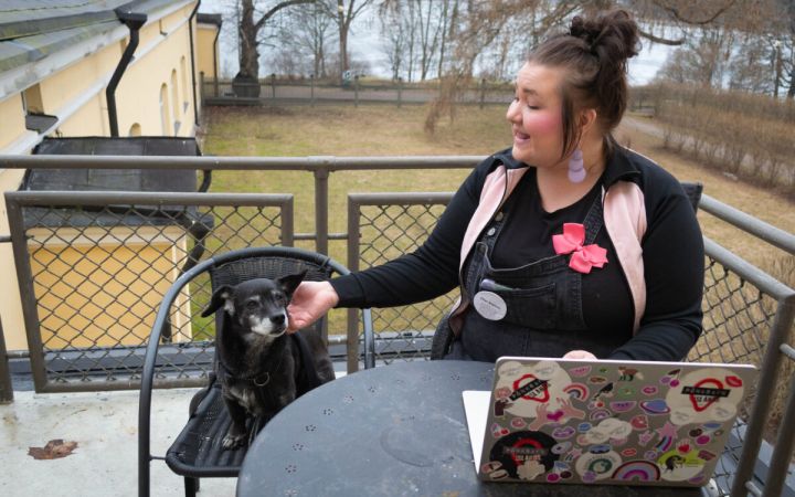 Nainen istuu parvelleella tietokoneen ääressä ja viereisesllä tuolilla on pieni musta koira.