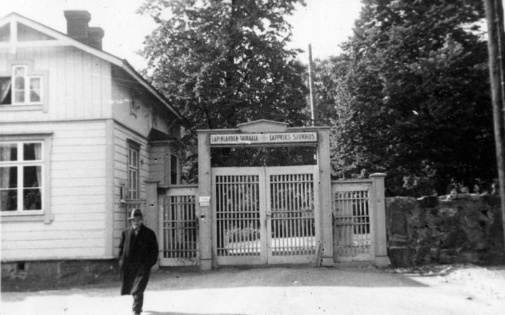 Mustavalkoisessa kuvassa mies kävelee poispäin sairaalan portilta. Portin vieressä on puinen rakennus.