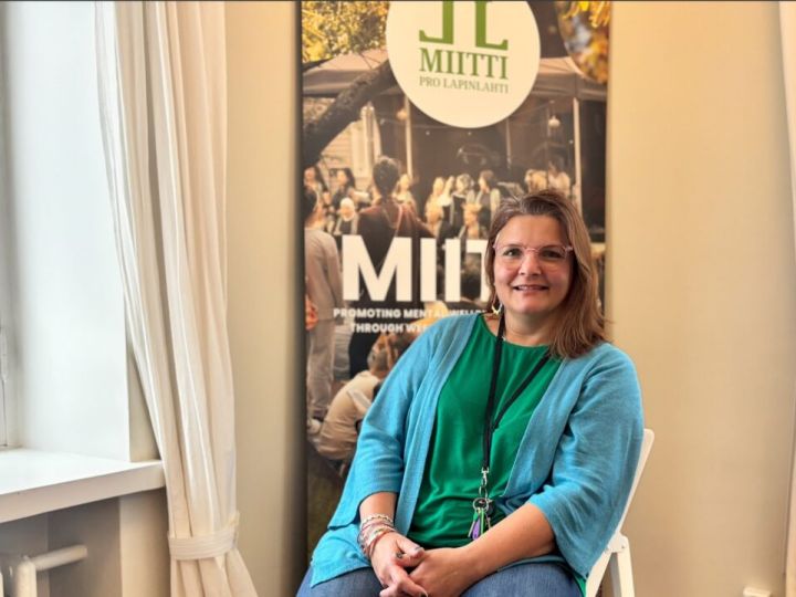 Nainen istuu MIITTI-julisteen edessä tuolissa ja hymyilee