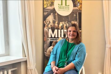 Nainen istuu MIITTI-julisteen edessä tuolissa ja hymyilee