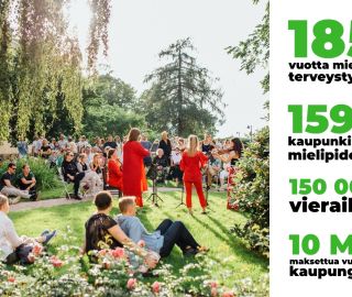 Joukko ihmisiä on kokoontunut aurinkoiseen puutarhaan kuuntelemaan viulutrion konserttia.