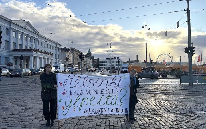 Kaksi ihmistä kannattelee banderollia Helsingin kaupungintalon edustalla auringon noustessa.
