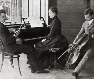 Mustavalkoinen kuva Sibeliuksen sisaruksista. Jean istuu vasemmalla sylissään viulu, Linda keskellä pianon ääressä ja Christian Sibelius oikealla sellon kanssa.