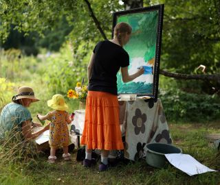 Kolme ihmistä puutarhassa, lapsi ojentaa maassa istuvalle maalia. Seisova nainen maalaa taulua.