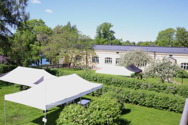 Kuvassa on puutarhapiha päärakennuksen toisen kerroksen parvekkeelta kuvattuna. Etualalla on pensaita ja katoksia ja taka-alalla talon pohjoissiipi.